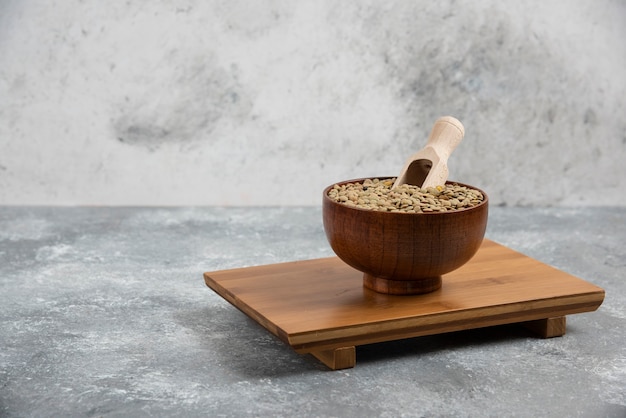 A small wooden bowl of dried seeds with a scoop, standing on a wooden board against a grey wall